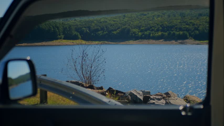 Scenic view of Colebrook River Lake in Connecticut, USA, seen through a car window with a rocky shore, roadside guardrail, and forested hills.