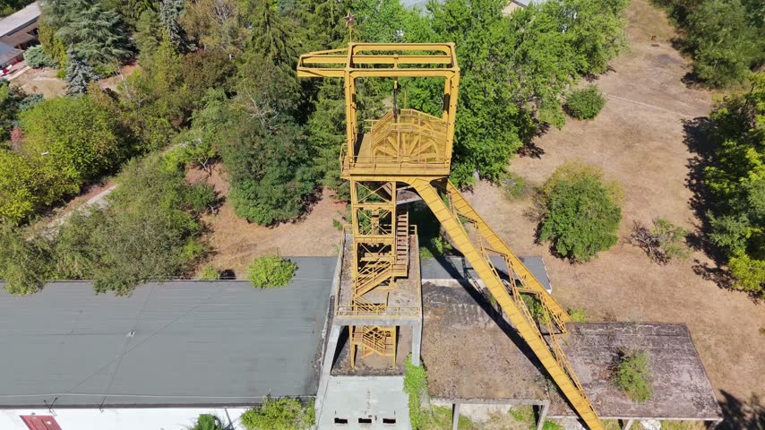 Aerial view of the historic Tiribesi-Akna coal mine headframe in Bátonyterenye, Hungary, standing above former industrial buildings amid summer trees and the Mátra foothills.