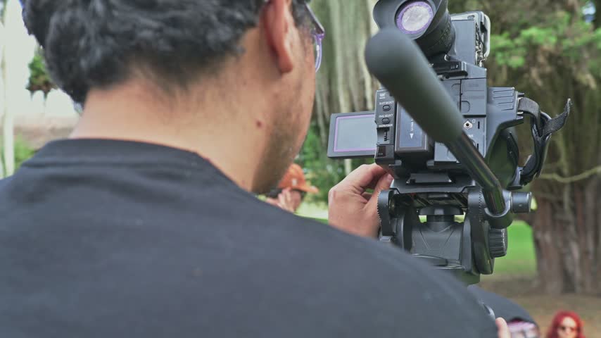 A cameraman operates a professional shoulder-mounted video camera on a tripod in a park, adjusting settings and focusing while capturing footage for an outdoor event, documentary or media production.