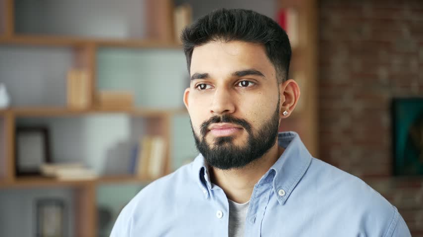 Close-up portrait of a handsome serious bearded man looking at the camera as he stands indoors. A confident businessman stays focused, posing calmly in his modern home office with a thoughtful look.