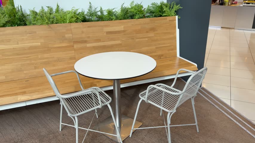 White round table and two light metal wire chairs are arranged next to a long wooden bench inside a modern mall food court. Indoor cafe seating, shopping center dining area, restaurant lounge zone.