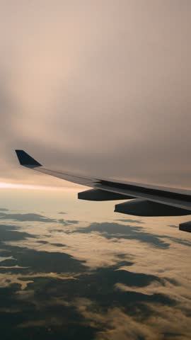 Vertical view of an airliner wing over low white clouds and patches of land under a grey morning sky on the final approach.