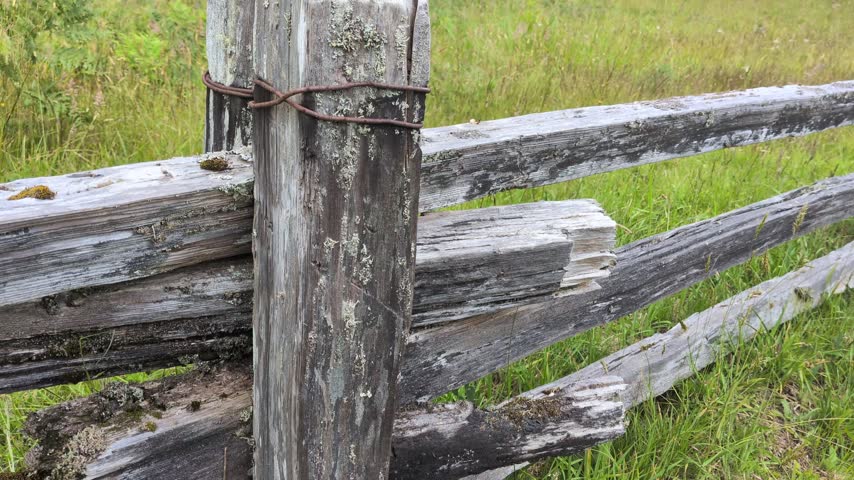 Close-up detailed view of a rustic weathered wooden split-rail fence post bound with wire in a lush field.