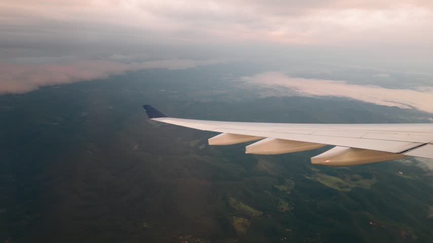 Airliner wing under a heavy overcast sky above dark forested hills and the edge of a low cloud layer tinted pink by the dawn light.