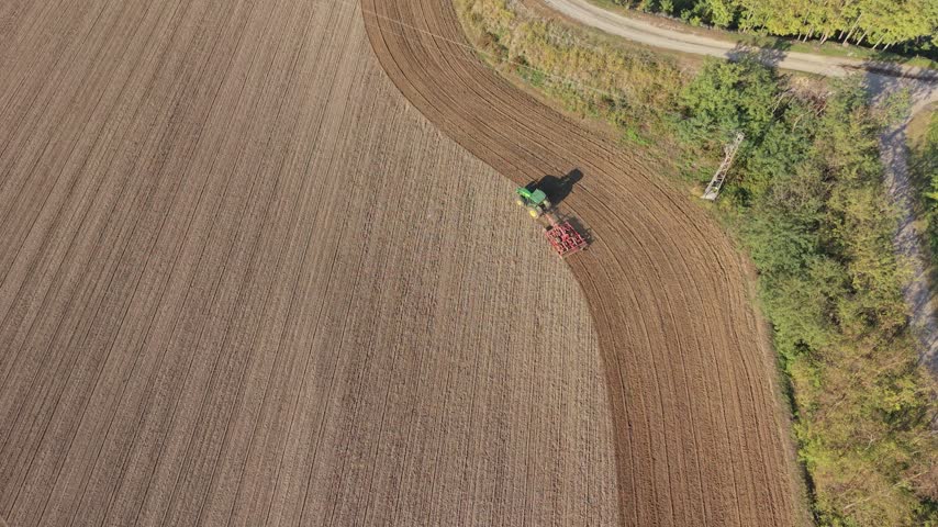 Aerial view of a green tractor plowing a brown field during autumn with nearby trees and a dirt road in Milan, Lombardy, Italy.
