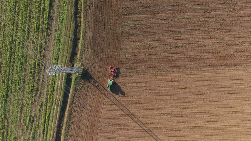 Aerial view of a green tractor plowing a brown field with a power line nearby in Milan, Lombardy, Italy.