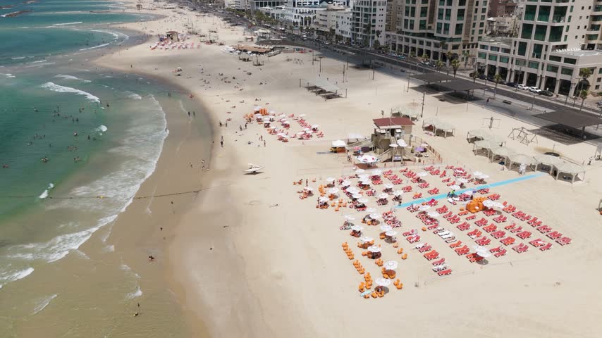Aerial view of the sandy beach in Tel Aviv with rows of sun loungers and umbrellas, clear turquoise water, and modern buildings along the promenade in Tel Aviv-Yafo, Tel Aviv District, Israel.