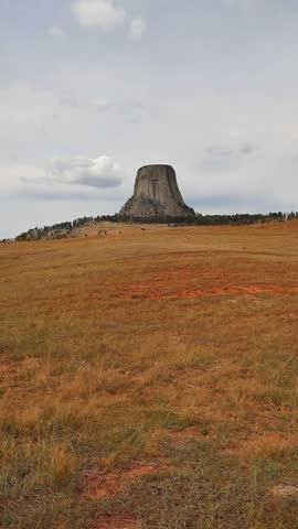 Vertical shot of Devils Tower National Monument standing above a windswept golden prairie with red soil and pine-covered hills under a cloudy sky, Wyoming.