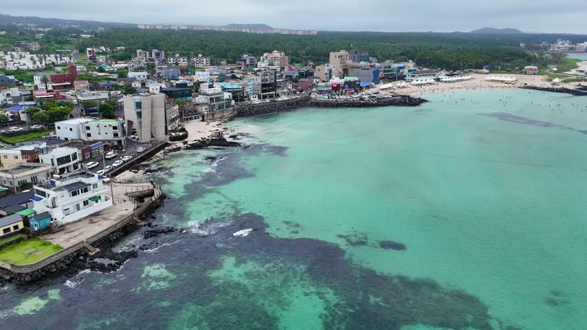 Aerial view of Hyeopjae Beach with turquoise waters, rocky coastline, and surrounding urban buildings in Cheju, Jeju-do, South Korea.