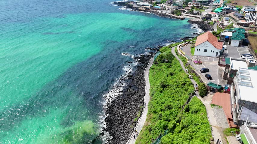 Aerial view of the coastline with turquoise water, volcanic rock cliffs, a coastal pathway, and buildings in Cheju, Jeju-do, South Korea.