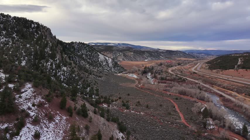 Aerial view of the Eagle River, walking trails, and Interstate 70 with snow-dusted mountains under a cloudy sky in Eagle, Colorado, United States.