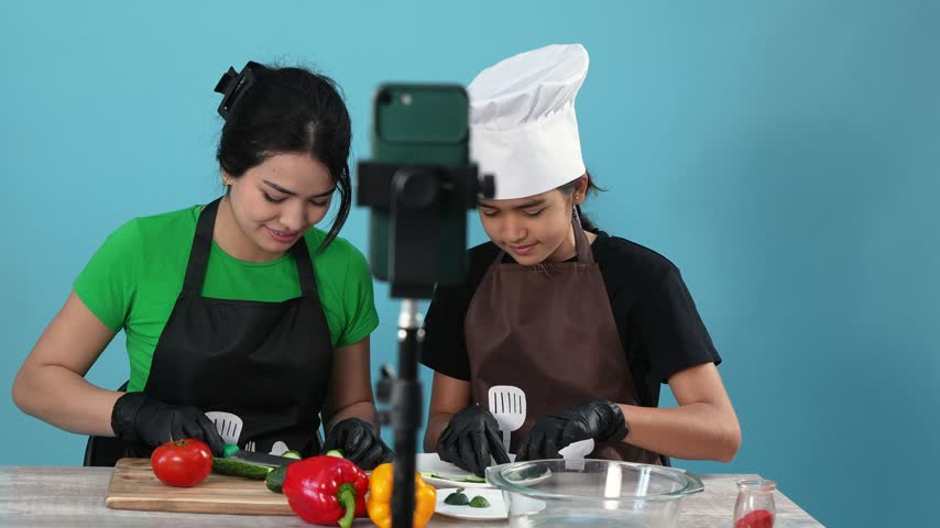 A woman and a young girl wearing a chef hat funnily eat cucumber slices while recording a cooking video blog on a smartphone against a blue background.