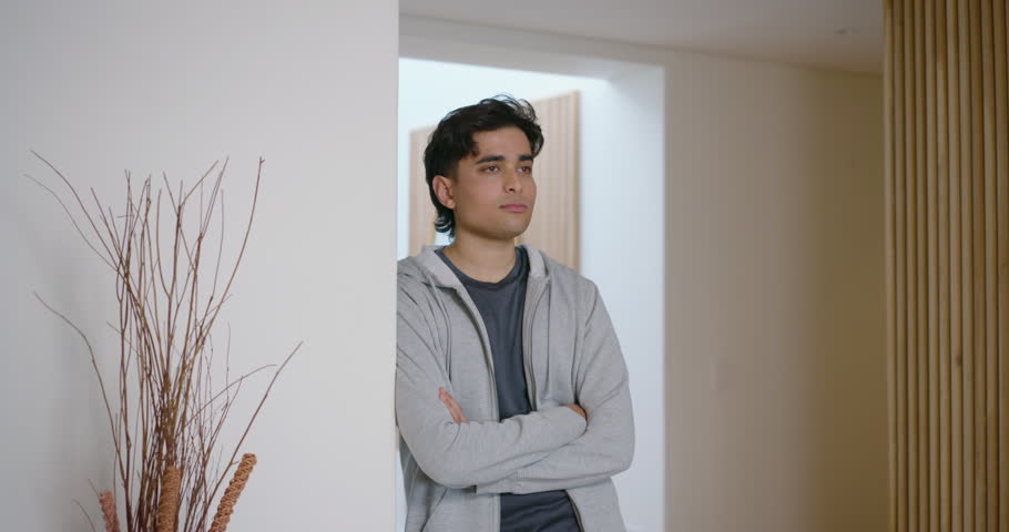 Asian man standing in entryway wearing hoodie, noticing off-screen guest, smiling by mirror. Portrait, interior, modern, minimalist, doorway, wooden, slatted