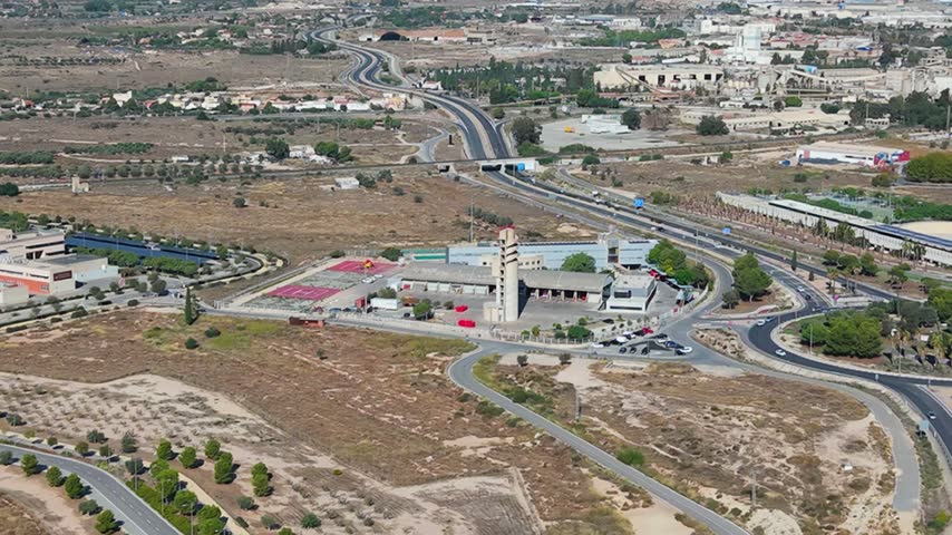 Aerial view of a fire station with a tall tower and red vehicles situated near a highway and roundabout in the dry landscape of Alicante, Valencian Community, Spain.