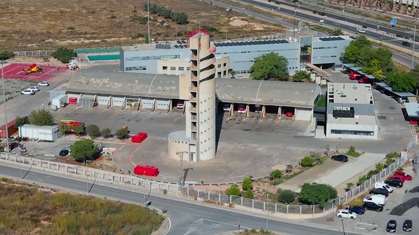 Aerial view of the fire station with its distinctive training tower, fire trucks parked in bays, and surrounding parking areas in Alicante, Valencian Community, Spain.