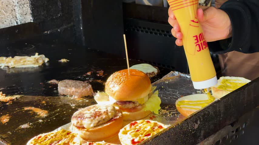 Close up view of cook squeezing mustard and mayonnaise sauce onto toasted bread rolls while preparing delicious homemade burgers at food stall.