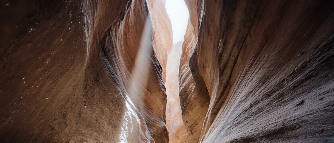 Golden sunlight streams down inside a deep narrow geological rock gorge. Smooth orange sandstone formations rise vertically toward an open distant sky. Natural warm light rays pierce through dusty air within this scenic remote cavern. Low angle upward perspective emphasizes massive sheer cliffs displaying rich earthy textures.