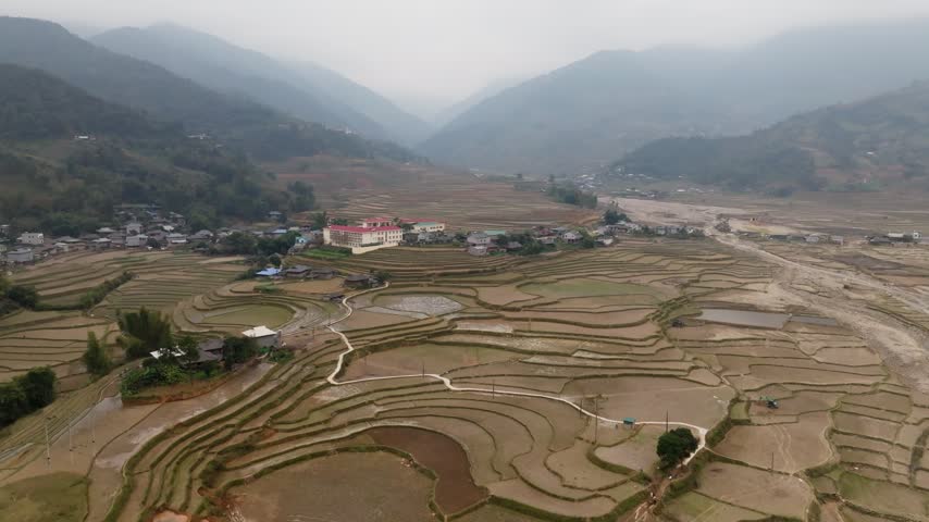 Drone rises and moves toward a village with a red-roofed school among fallow and flooded terraces in Tú Lệ valley, northern Vietnam.