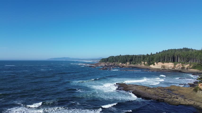 Aerial view of rugged Oregon coast near Depoe Bay with waves crashing on rocks and forested headland, drone flying forward