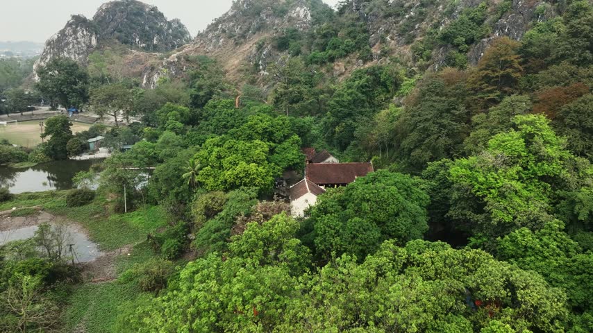 Drone approaches a temple with dark tiled roofs and white walls among lush green trees on the rocky limestone hillside of Chùa Trầm, Vietnam.