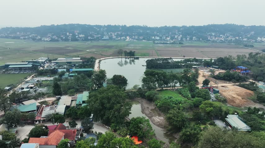 Aerial flight toward a pond near Chùa Trầm pagoda in Chương Mỹ, Hanoi, with village rooftops, trees, rice fields and low hills on the hazy horizon, Vietnam.