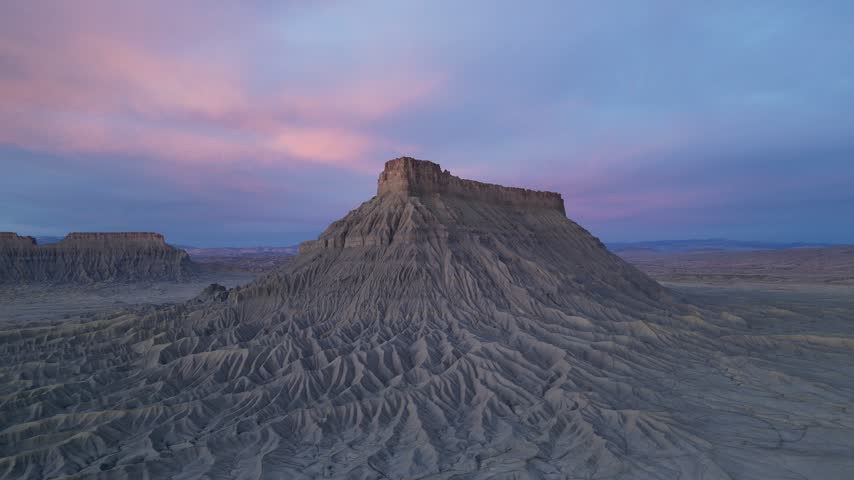 Aerial view of Factory Butte surrounded by eroded badlands and desert terrain under a soft sunset sky in Wayne County, Utah, United States.