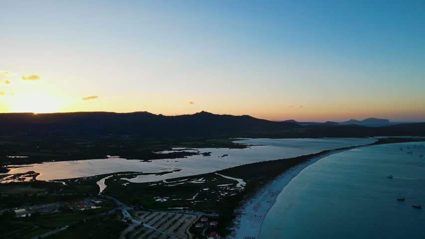 A stunning aerial perspective of the Sardinian coast at dusk, featuring a long sandy beach, calm lagoon waters, and silhouettes of mountains under a vibrant sunset sky