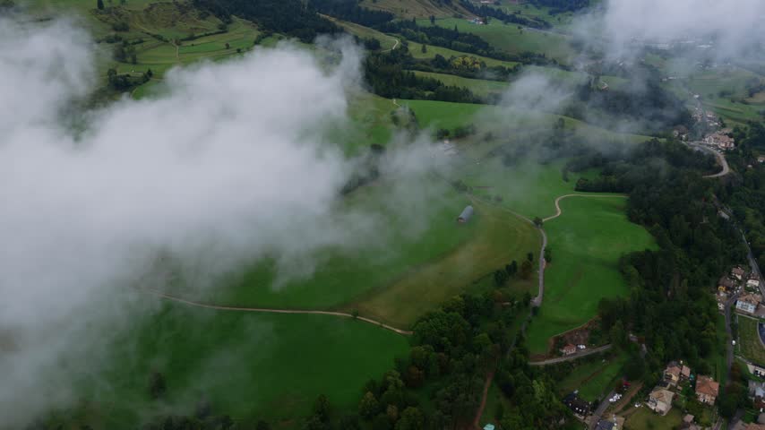 The camera crosses a cloud and comes out over mown alpine meadows and farm tracks, a rocky ridge standing behind the forest. Aerial fly-through, tilt up as the ridge appears.