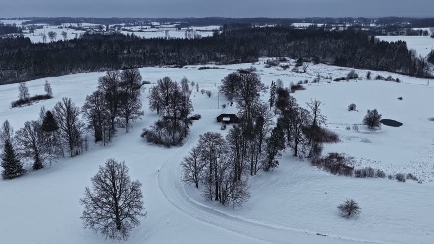 Aerial view of a small house nestled among snow-covered trees and fields in a rural winter landscape in Latvia.
