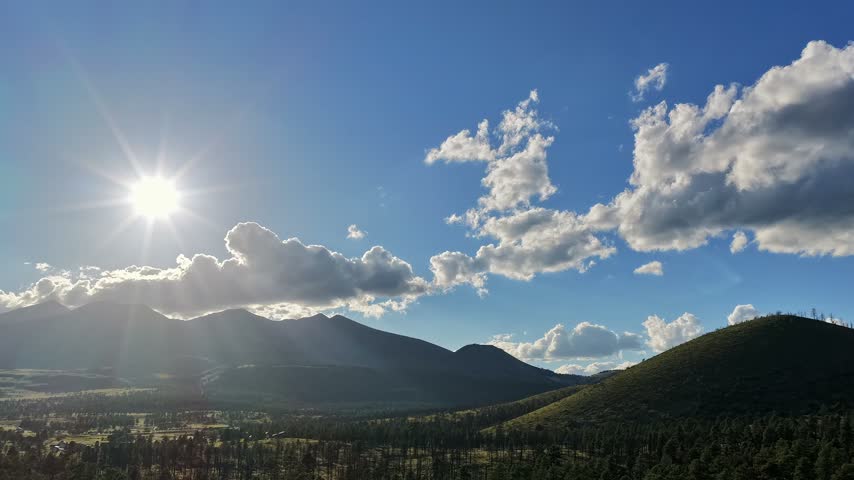 4K drone time lapse of the bright afternoon sun shining over the San Francisco Peaks in Flagstaff, Arizona. Scattered white clouds drift above the forested volcanic landscape.