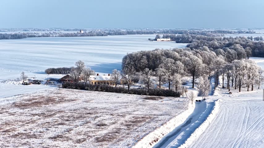 Aerial view of a small building nestled among frost-covered trees in a vast snow-covered rural landscape, Latvia.