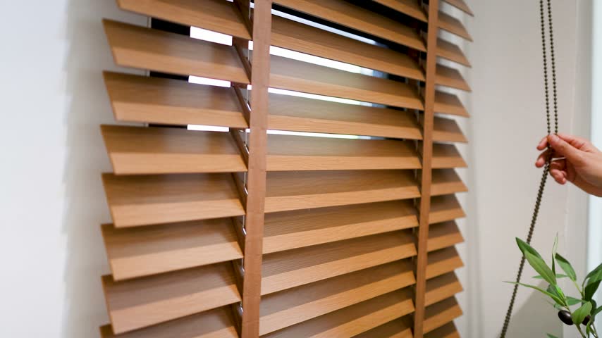 Person hand pulling beaded chain to tilt wooden venetian blinds, adjusting sunlight entering through window in minimalist room.