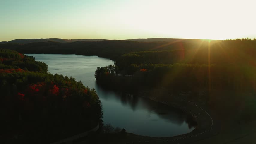 The sun peeks over the horizon at Barkhamsted Lake in Connecticut surrounded by warm fall foliage at dusk