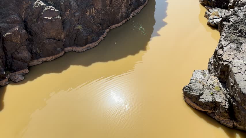 Muddy water pool with ripples surrounded by steep rocky canyon cliffs and bright sun reflection.
