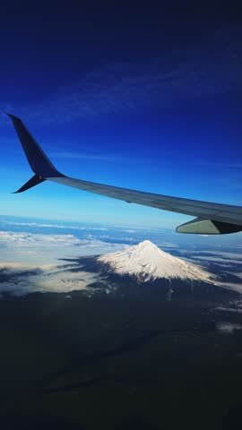 Vertical aerial shot from outside an airplane window of the stunning snow-capped Mount Hood, Oregon, rising behind the wing, with Cascade Range peaks stretching into the distance under a blue sky