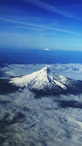 Vertical aerial shot from an airplane window of the large, stunning snow-capped Mount Hood in Oregon, with Cascade Range peaks in the distance under a bright, sunny blue sky, flying away from Portland