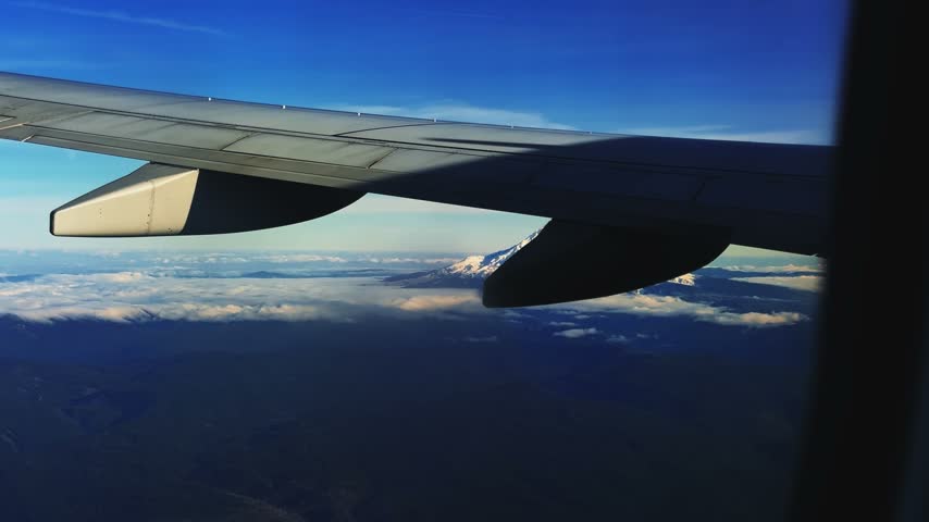 Aerial shot from outside an airplane window of the stunning snow-capped Mount Hood, Oregon, rising behind the wing, with Cascade Range peaks stretching into the distance under a clear, sunny blue sky.