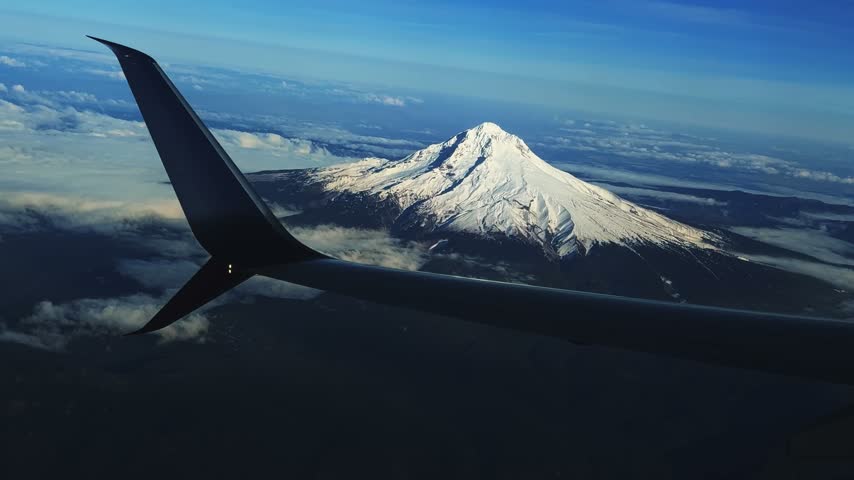 Aerial shot from outside an airplane window of the stunning snow-capped Mount Hood, Oregon, rising behind the wing, with Cascade Range peaks stretching into the distance under a clear, sunny blue sky.