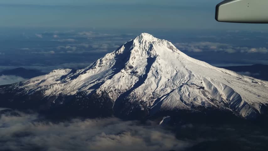 Aerial shot from an airplane window of the large, stunning snow-capped Mount Hood in Oregon, with Cascade Range peaks in the distance under a bright, clear sunny blue sky, flying away from Portland.