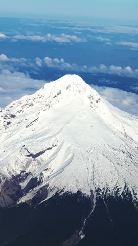 Vertical close up aerial shot from an airplane window of the large, stunning snow-capped Mount Hood in Oregon, with Cascade Range peaks in the distance under a bright, sunny blue sky