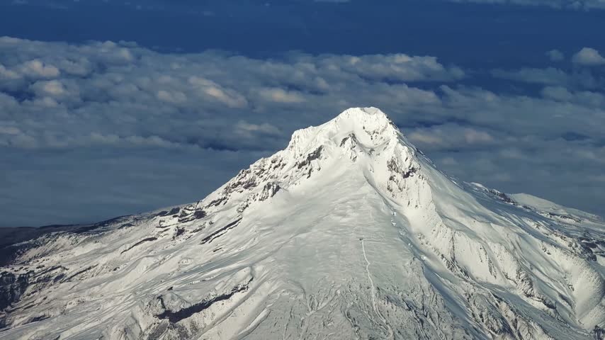 Close up aerial shot from an airplane window of the large, stunning snow-capped Mount Hood in Oregon, with Cascade Range peaks in the distance under a bright, sunny blue sky