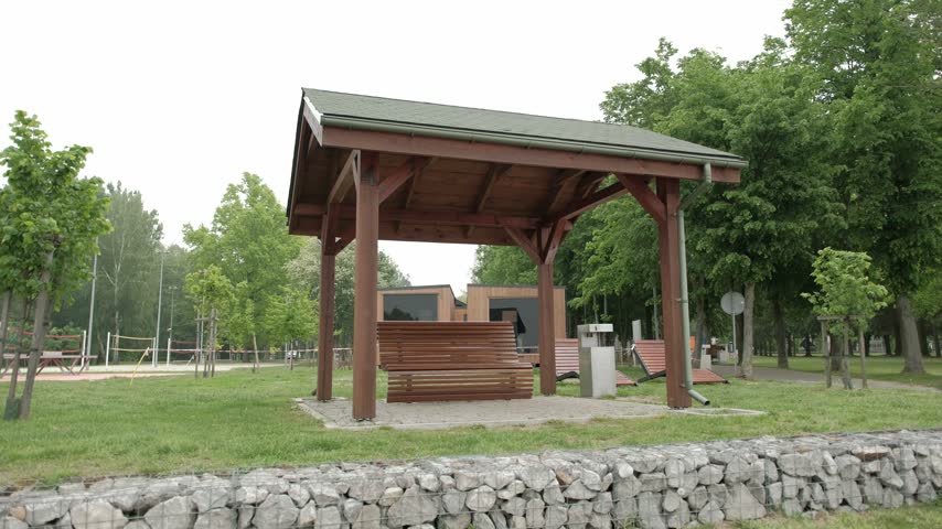 A wooden gazebo sheltering a bench in a green public park with trees and recreational facilities