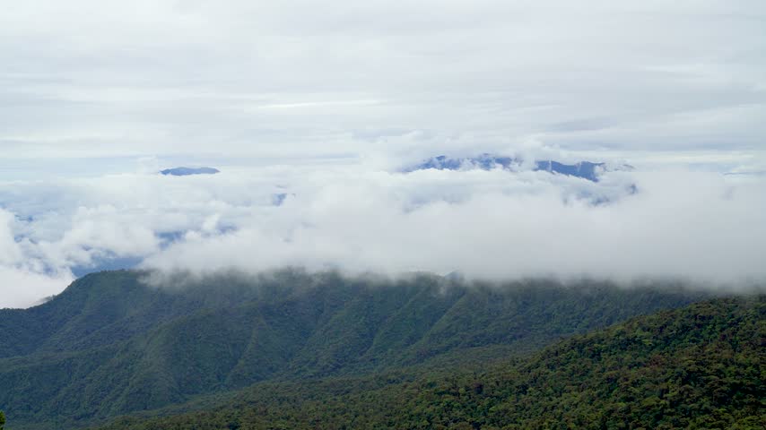 Poas Volcano National Park in Alajuela, Costa Rica, clouds drifting over green rainforest-covered mountains as distant volcanic peaks emerge above the cloud layer