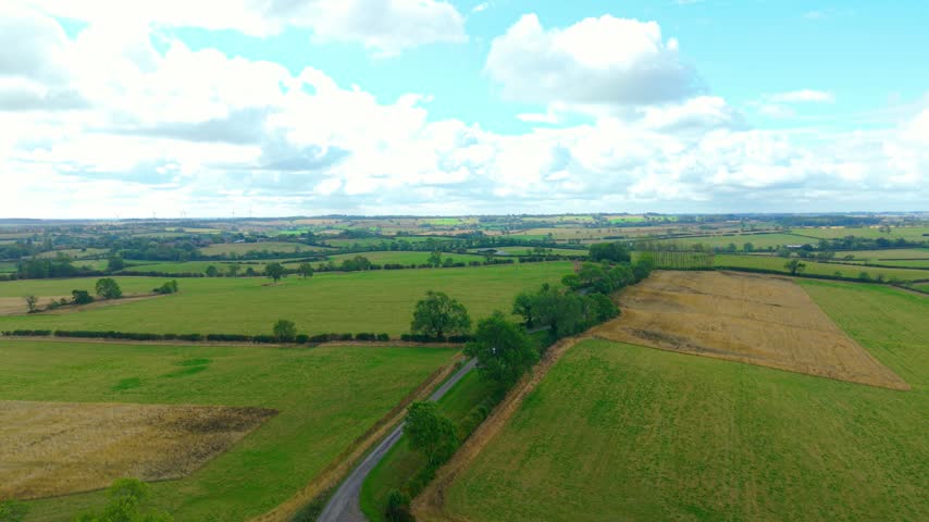 Agricultural countryside with green fields, country road and scattered trees. Rural landscape with cultivated farmland beneath bright cloudy sky. Aerial view across patchwork fields with distant wind