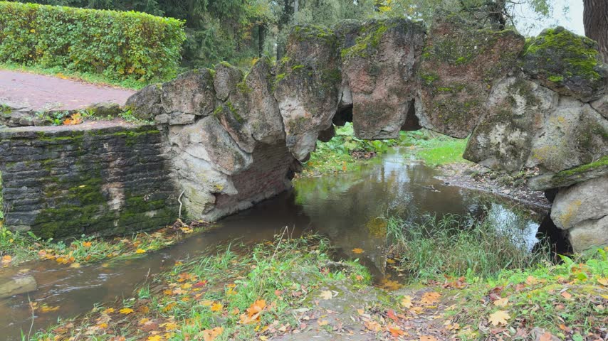 a stone bridge in a park, which has begun to become overgrown with wild vegetation, and a stream flowing beneath it
