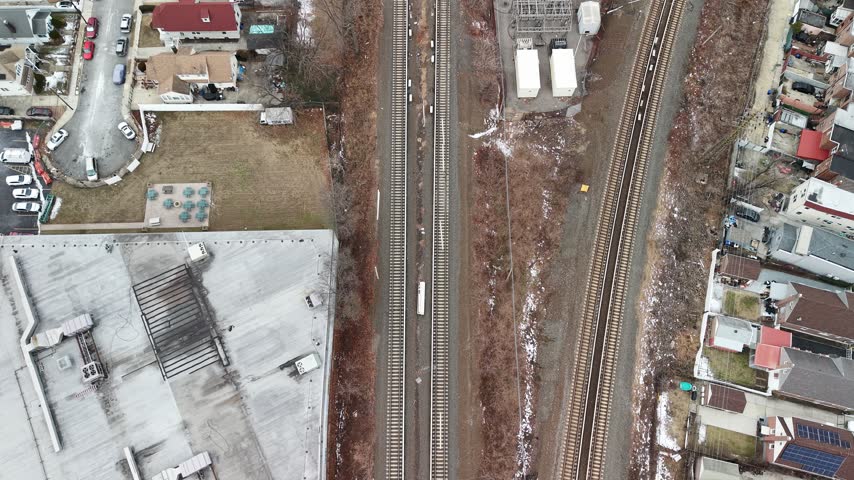 An aerial glide follows railway tracks beside homes and parking lots in Queens. Parallel tracks divide compact residential blocks while the camera moves along the corridor under a pale winter sky.
