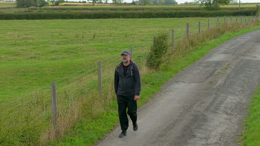 Rural hiker walking beside wire fence and green pasture on quiet country lane. Bearded middle aged man with backpack traveling through farmland. Countryside tourism and outdoor recreation across