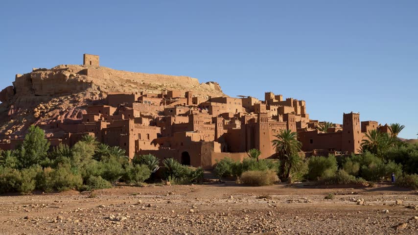 Ait Ben Haddou near Ouarzazate, Morocco, the fortified mud brick ksar rising on a hillside with towers, earthen houses and a granary on top, palm trees and shrubs in front