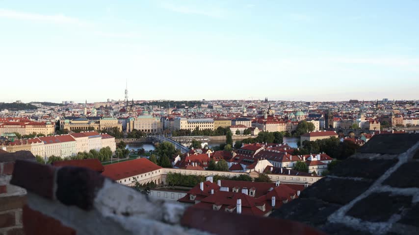 Prague panoramic skyline overlooks the Vltava River and historic bridges as red rooftops and famous church towers stretch across the Czech capital beneath a pale blue sky