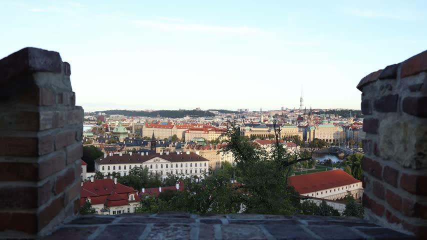 Prague city panorama framed by ancient brick walls overlooking historic rooftops elegant towers the Vltava River and bridges with lush green trees beneath a pale blue sky
